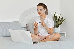 Sad upset young adult woman wearing white t shirt and shorts sitting on bed in front of laptop holding pregnancy test with