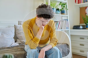 Sad upset young man sitting on couch at home, touching his head with hands