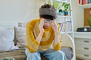 Sad upset young man sitting on couch at home, touching his head with hands