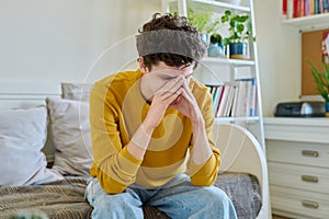 Sad upset young man sitting on couch at home, touching his head with hands