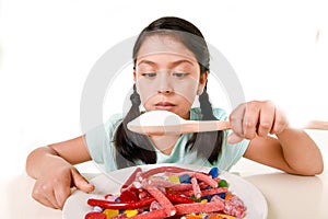 Sad and vulnerable hispanic female child eating dish full of candy and gummies holding sugar spoon in wrong diet concept