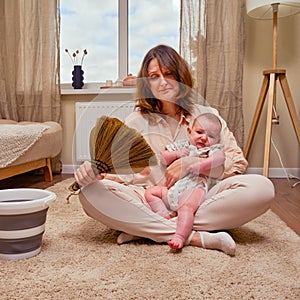 A sad woman with a baby is sitting on the floor in a home living room, problems with household chores at the birth of a child