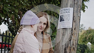 A sad woman with a child stands at a pole where an advertisement for a missing young woman has been announced. Search