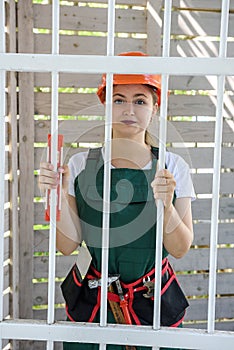 Sad woman in coverall standing behind closed gate. She is tired and wearing helmet and tool belt