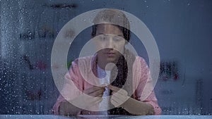 Sad woman holding engagement ring behind rainy window, upset after break up