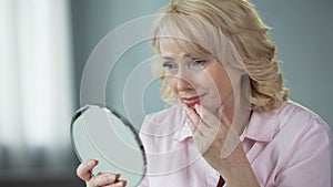 Sad woman looking at face reflection and crying, unhappy with wrinkled skin