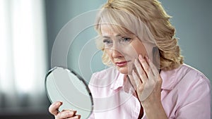 Sad woman looking at face reflection and crying, unhappy with wrinkled skin