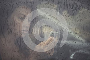 Sad woman looking in the mobile phone and reading message in the window with rain drop in the car.