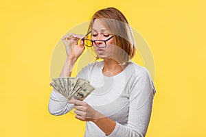 A sad woman looks through her glasses at a small fan of dollars in her hands. Yellow background. The concept of finance and