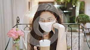 Sad woman in medical mask sitting at the table at cafe or restaurant and looking at cocktail and sigh. Social distancing