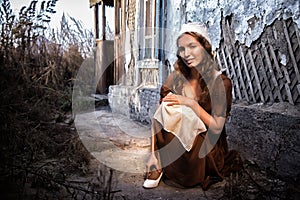 Sad woman in a rustic dress sitting near old brick wall in old house and trying to dress a white shoe. Cinderella style