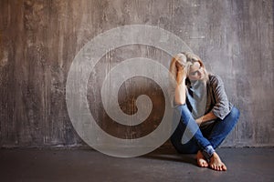 Sad woman sits on the floor. Depression and chronic fatigue. Young beautiful blonde in a gray sweater and jeans