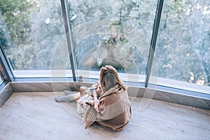 Sad woman sitting on a warm floor in a socks wrapped in a woolen blanket near the large window in light. autumn mood, warmth and