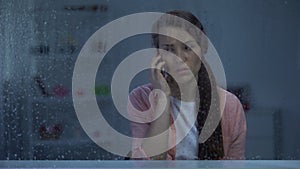 Sad woman talking phone behind rainy window, shocked by bad news from family