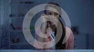 Sad woman talking phone behind rainy window, shocked by bad news from family