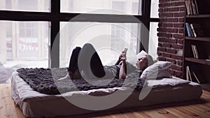 Sad woman using smartphone, lying on bed by window, overlooking city street