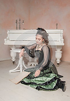 Sad woman wearing medieval vintage Victorian Style dress sitting on the floor near piano with letter