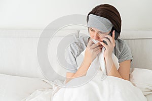 Sad woman wearing sleeping mask using smartphone as she sits in bed covered with duvet