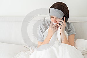 Sad woman wearing sleeping mask using smartphone as she sits in bed covered with duvet