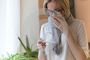 Sad woman wrapped in scarf feeling flu symptom, looking at thermometer. Virus, cold symptom