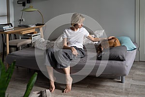Sad worried middle-aged woman sitting on sofa caring for old sick pet at home, depression in dogs