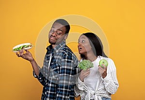 Sad young african american wife with broccoli and apple in hands looks at cheerful guy with sandwich