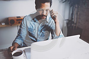 Sad young African man reading world news on laptop while sitting at the table a sunny morning.Concept of coworking