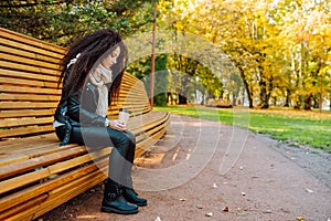 Sad young afro-haired woman sit alone on a wooden bench in autumn park at sunny warm day. Portrait of alone woman