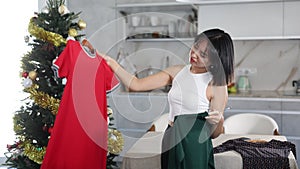 Sad young Asian girl trying to choose a dress standing next to Xmas tree in the kitchen