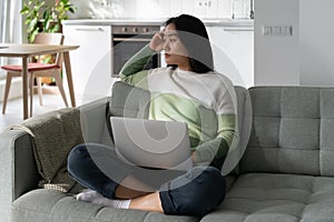 Sad young Asian woman freelancer tired sits on sofa with laptop on lap suffering lack of motivation