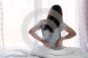 Sad young Asian woman touching back feeling backache morning discomfort low lumbar muscular kidney pain sit on bed after bad sleep