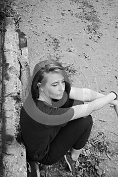 A sad young attractive red-haired girl is sitting on her haunches, leaning against an old sidewalk stone.