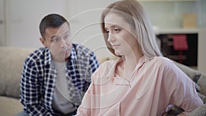 Sad young beautiful woman sitting on couch as blurred Caucasian man talking at background. Portrait of unhappy