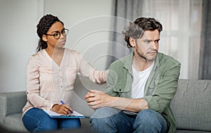 Sad young black lady doctor calms unhappy despaired caucasian male patient in office clinic interior