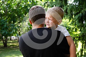 Sad young boy crying on fathers hands in nature, park, outdoor. Men holds his son, hugs and comforts. Family love, care and moral