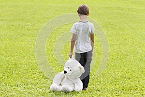 Sad young boy is holding a teddy bear and standing on the meadow. Child looking down. Back view. Sadness, fear