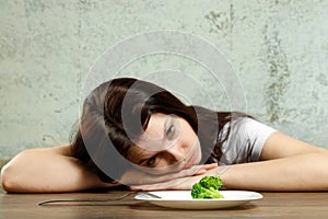 Sad young brunette woman dealing with anorexia nervosa or bulimia having small green vegetable on plate. Dieting problems, eating photo