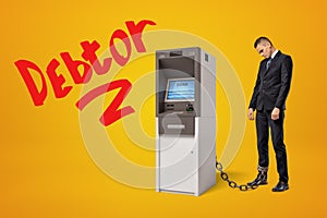 Sad young businessman standing in half-turn chained to ATM on amber background with big red title `Debtor`. photo