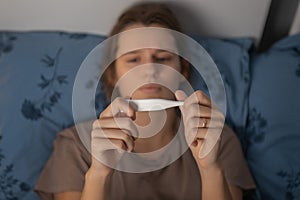 Sad young caucasian girl lying on pillow and looking on thermometer. Sickness woman hold temperature meter in hands.