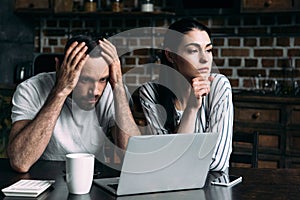 sad young couple sitting on kitchen with laptop