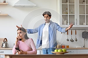 Sad young european woman with smartphone ignores offended angry screaming gesticulating husband photo