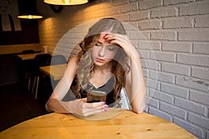 Sad young girl sitting in a cafe looking tired her phone, unhappy girl watching her black phone,