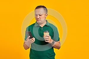 Sad young handsome man holding smartphone and cup of coffee on yellow background