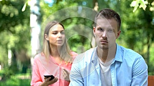 Sad young man looking camera, annoyed jealous girlfriend holding phone, distrust photo