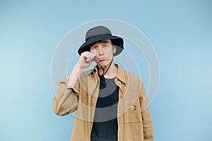 Sad young man in panama and shirt cries on a blue background and wipes tears. Isolated