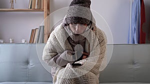 Sad young man sit at the table at freezing cooled flat in warm cap and blanket shiver tremble with cold, using smart