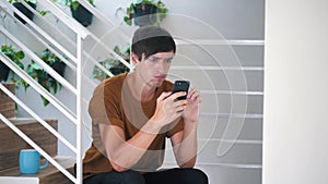 Sad young man sits on the stairs at home and uses a smartphone.