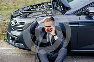 Sad young man use smartphone sitting on road near the broken car opened the hood, calling car assistance services