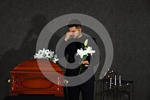Sad young man with white lilies near casket in funeral