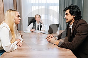 Sad young man and woman looking at each other angrily getting divorced sitting at table with layer in office